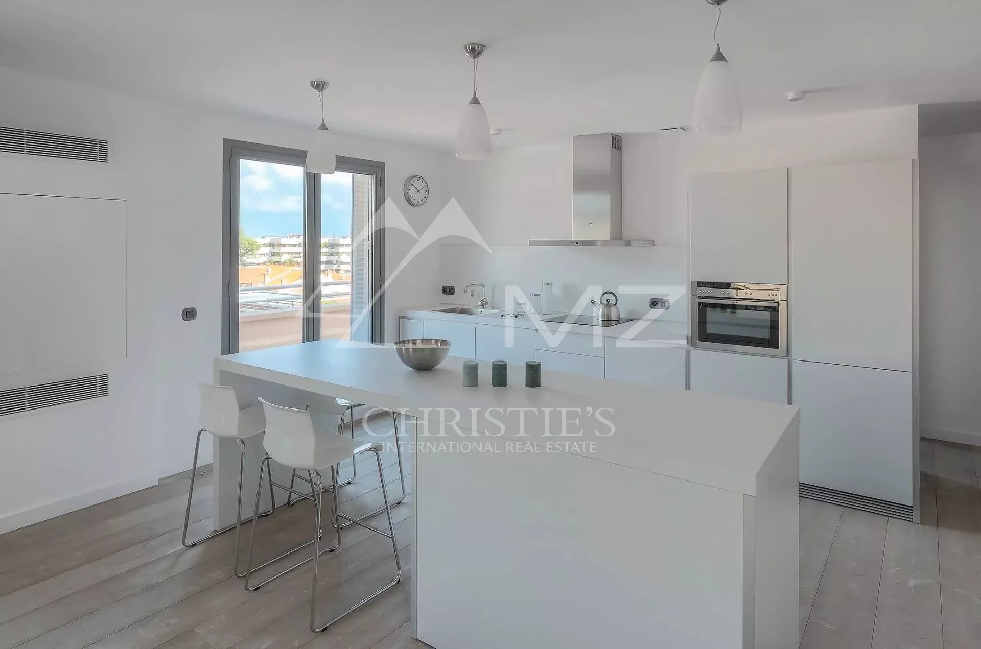 Bright white kitchen with a large central island, white bar stools, pendant lights, and a balcony door revealing an urban view.