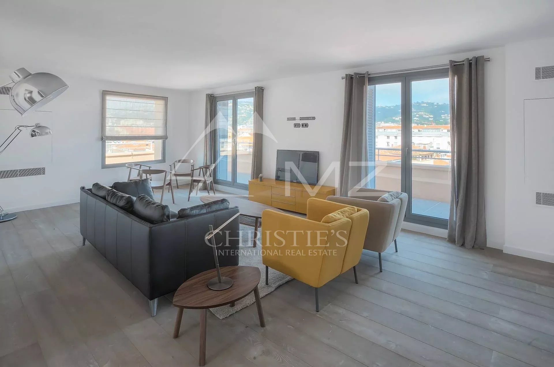 Spacious modern living room with a dark leather sofa, two mustard yellow chairs, a wooden coffee table, and a TV on a low console; large glass doors to a balcony and city views.