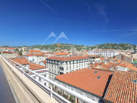 View of a Mediterranean-style town with orange-red tiled rooftops and white buildings under a bright blue sky, seen from a balcony railing edge on the left.