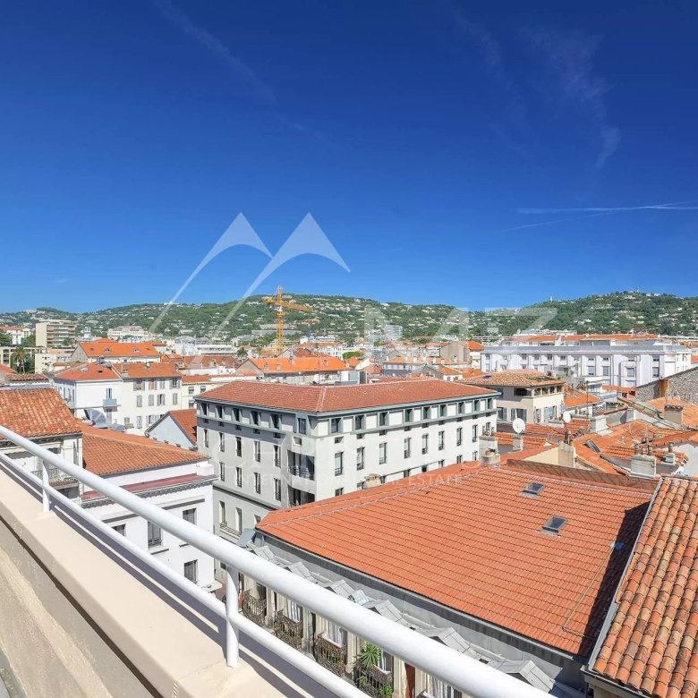 View of a Mediterranean-style town with orange-red tiled rooftops and white buildings under a bright blue sky, seen from a balcony railing edge on the left.