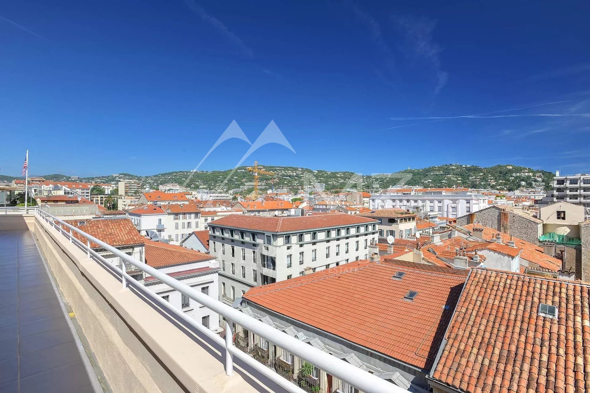 View of a Mediterranean-style town with orange-red tiled rooftops and white buildings under a bright blue sky, seen from a balcony railing edge on the left.