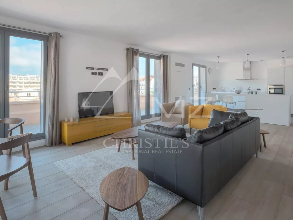 Open-plan living area with a black leather sofa facing a yellow TV console and a wall-mounted TV; large windows and balcony doors bring in natural light.