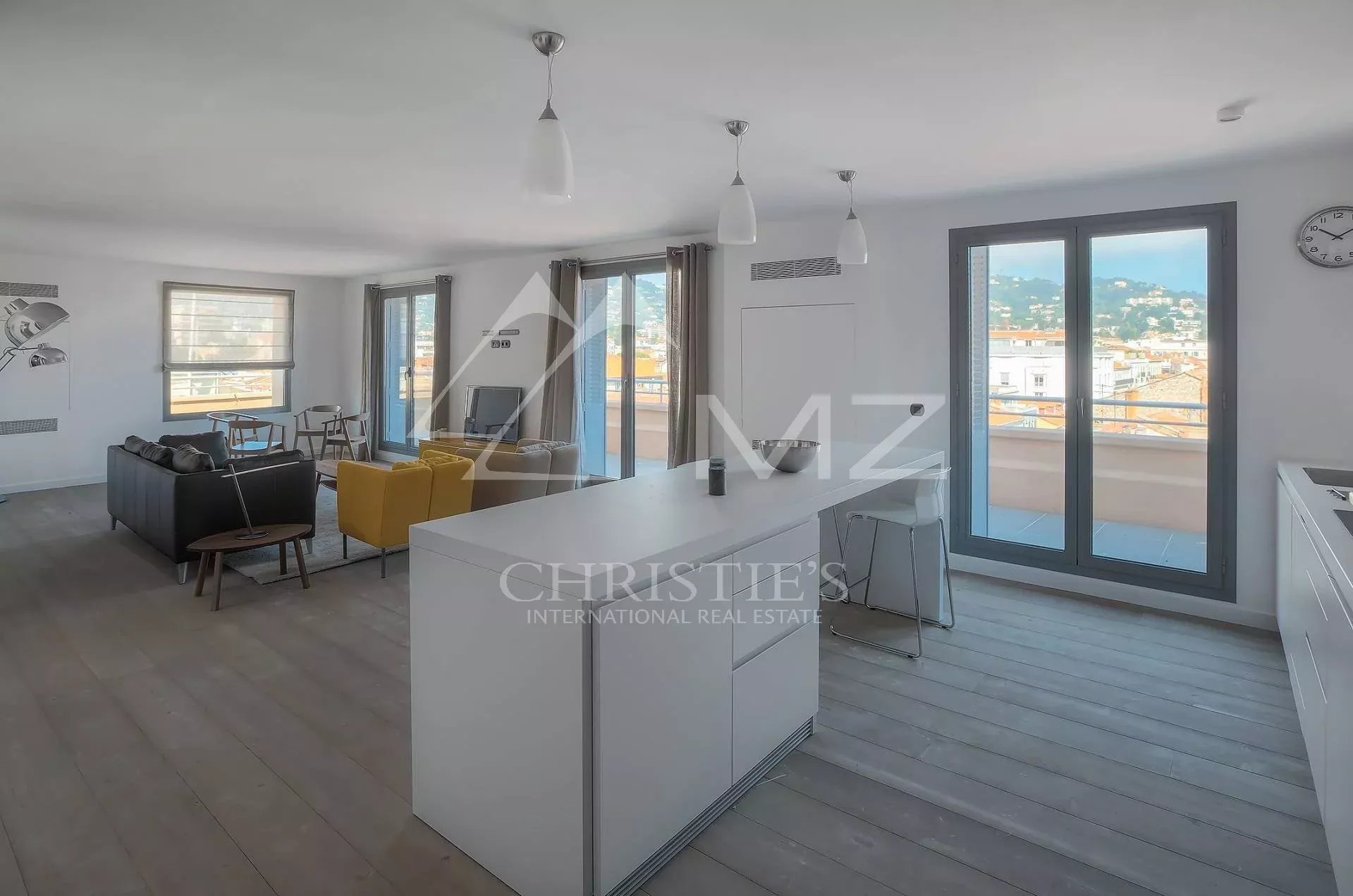 Open-plan living area with a white kitchen island, a yellow chair, and a gray sofa facing large windows and a balcony with a city view.