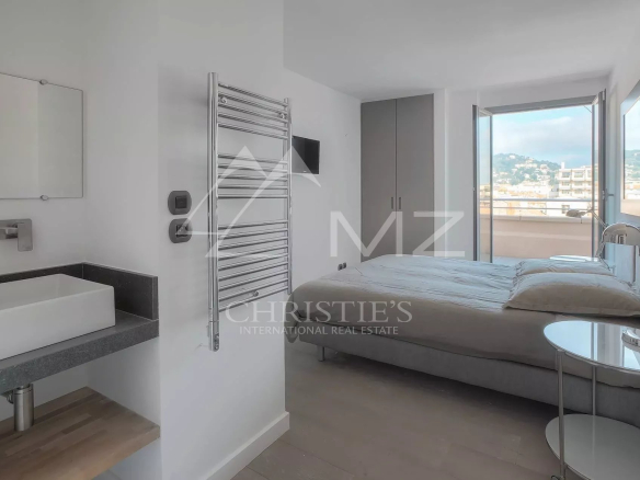 Hotel bedroom with a double bed, open balcony door showing cityscape and hills beyond, and a bedside table with a lamp.