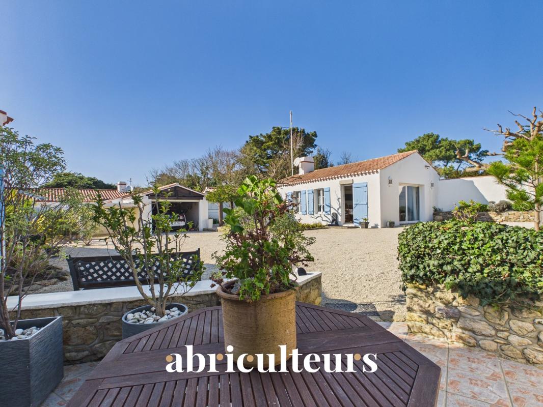 Sunny courtyard of a white Mediterranean villa with blue shutters and gravel yard, outdoor table in foreground.