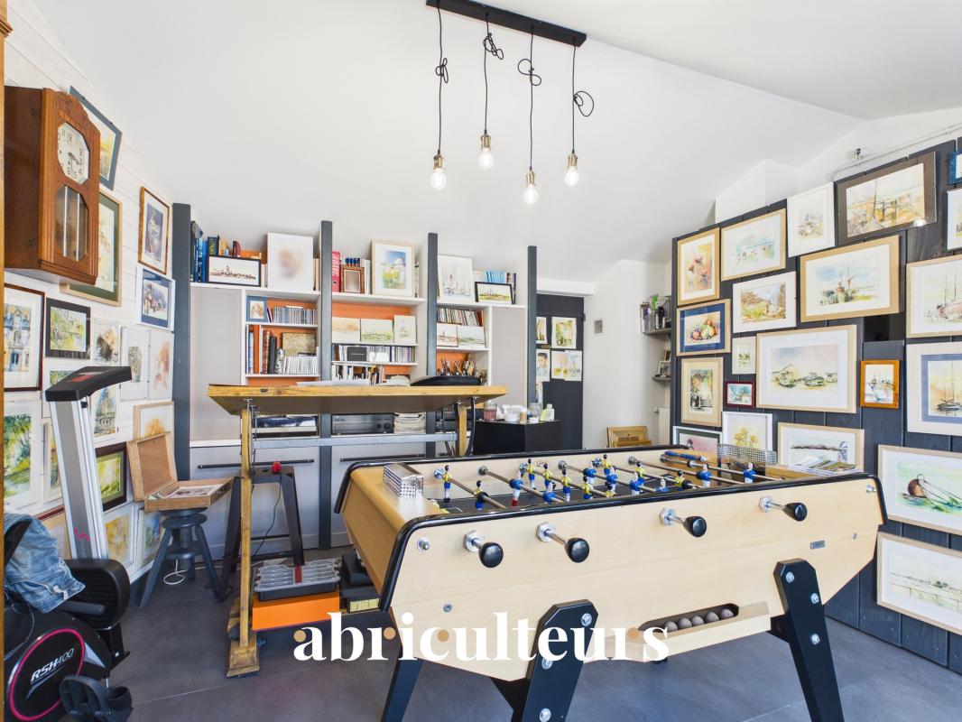 Art studio interior with a foosball table in the foreground, walls lined with framed paintings and drawings, and shelves of books and supplies behind.