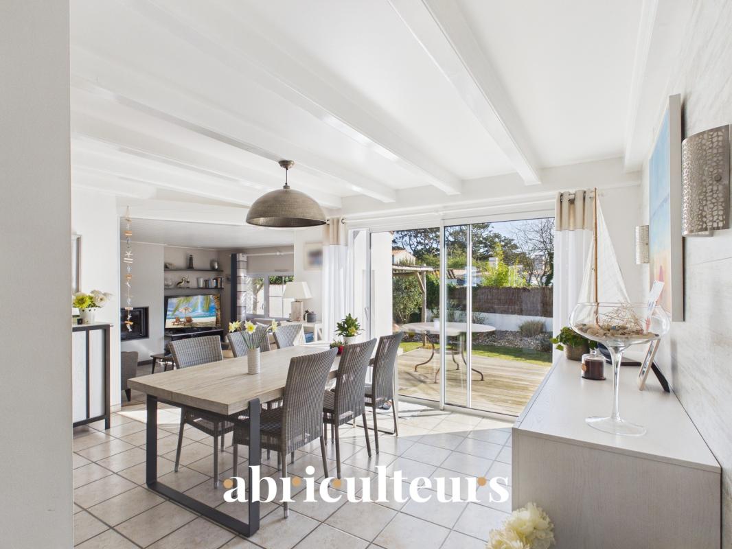 Bright, open-plan dining room with a long wooden table, wicker chairs, and large sliding glass doors to a sunny backyard patio.