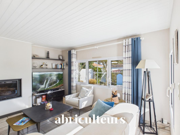 Bright living room with white seating, a flat-screen TV on a shelf, and a coffee table beneath a large window with blue-and-white curtains.