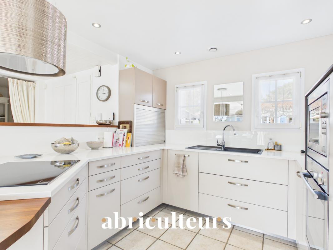 Bright white U-shaped kitchen with sleek cabinetry, double sink under windows, and stainless appliances; 'abriculteurs' watermark at bottom.
