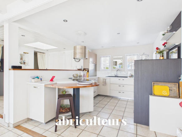 Bright, modern white kitchen with an island, wooden countertop, and stainless steel range hood; tiled floor and large windows behind the sink area.