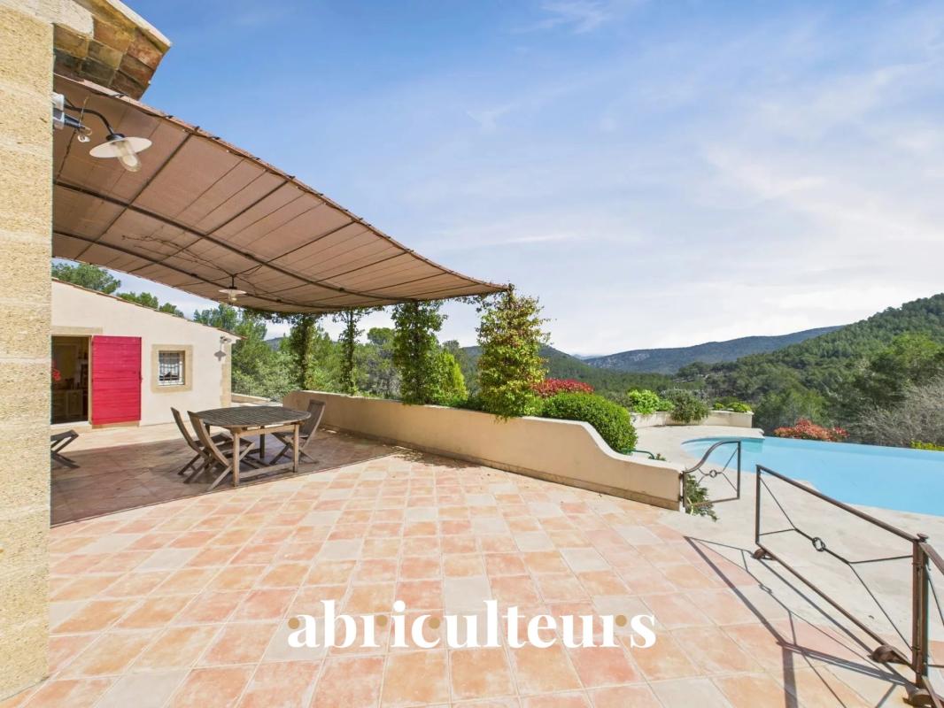 Outdoor tiled terrace with a wooden dining table and chairs under a fabric canopy, overlooking hills and a pool.