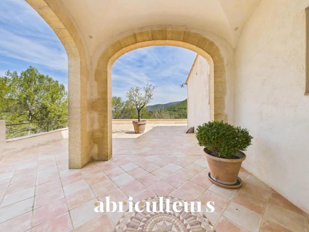Terrace with stone arches, pink-tiled floor, and a potted shrub; view of trees and hills beyond; watermark 'abriculteurs'.