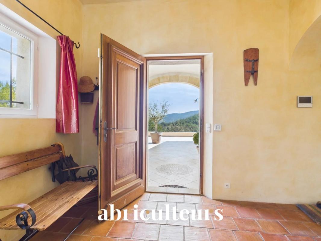 Sunlit entry hall with warm yellow walls, open wooden door to a tiled patio and distant hills, and a wooden bench against the left wall.