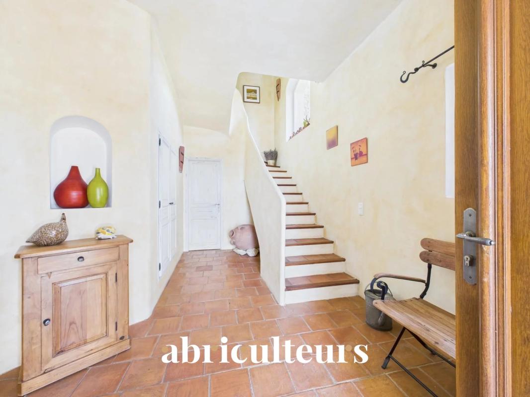 Warm home entry hall with terracotta floor, a wooden cabinet to the left, two ceramic vases in a wall niche, a wooden bench by the wall, and a curved staircase upward; 'abiculteurs' text appears at the bottom.