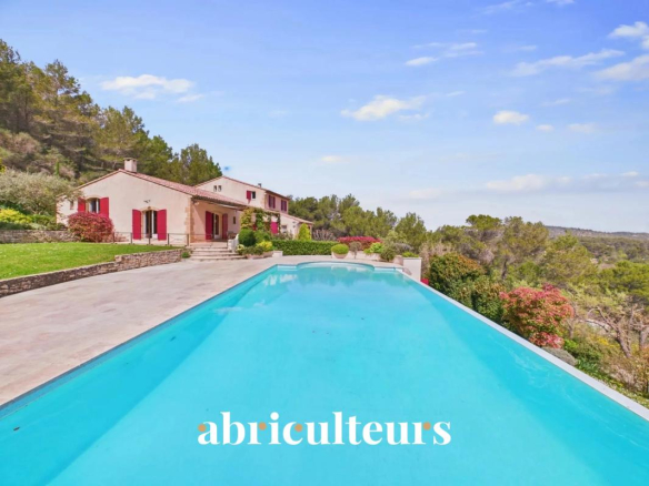 Villa with red shutters overlooking a hillside, with a long turquoise pool in the foreground under a blue sky.