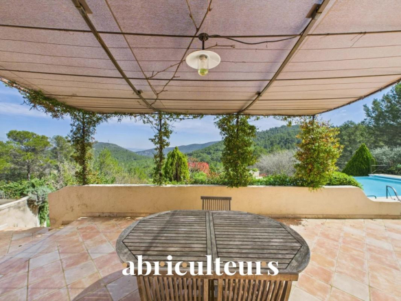 Patio scene with a wooden round table under a beige canopy, overlooking a lush garden and distant mountains; the word 'abriculteurs' appears at the bottom.