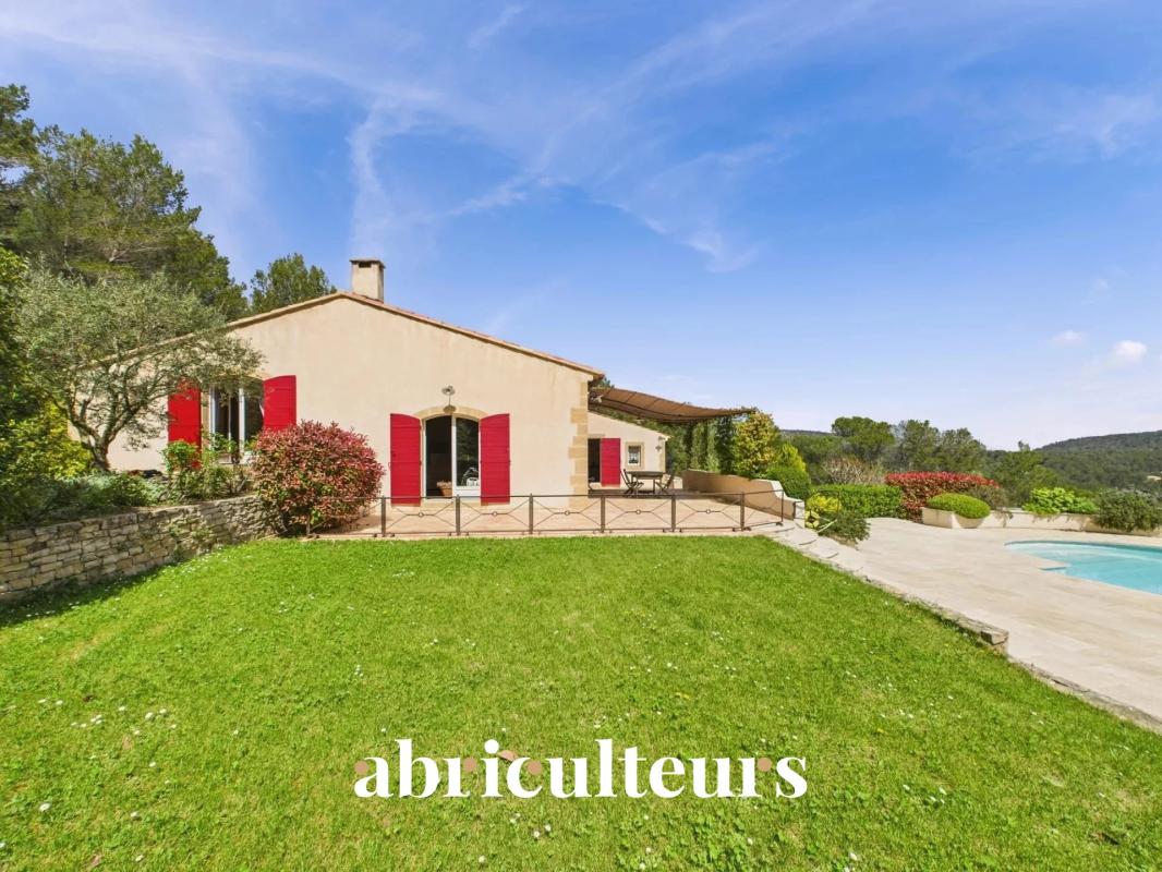 Villa with cream walls and red shutters, green lawn, stone wall, and a pool on a sunny hillside.