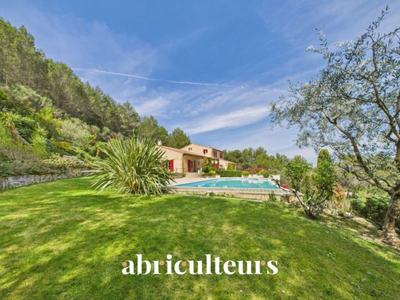 Sunlit hillside garden with a rectangular swimming pool, beige house with red shutters, and lush trees against a bright blue sky.