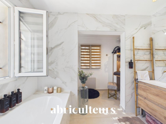 Bright marble bathroom with an open window, a white tub with dark toiletries, and a doorway leading to a dressing area with a plant and a wooden ladder