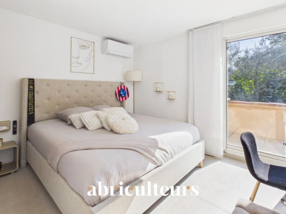 Sunlit minimalist bedroom with a large beige upholstered bed, neutral bedding and pillows, wall art, and a sliding glass door to a balcony.