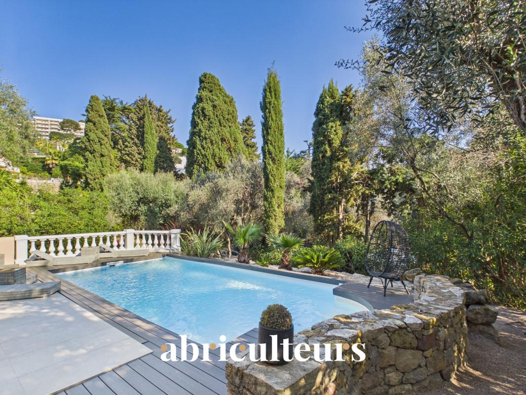 Luxurious backyard with a rectangular pool, wooden deck, stone wall, and tall cypress trees under a clear blue sky, with a logo reading 'abriculteurs' at the bottom.