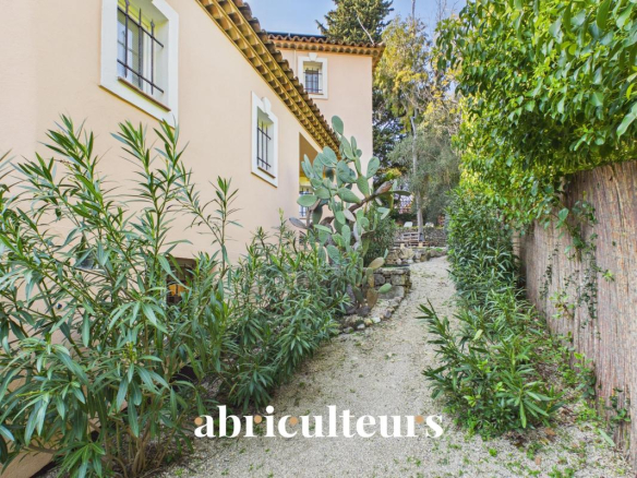 Garden path between a beige house and a wooden fence, with tall green shrubs and cacti along both sides; watermark 'abriculteurs' at the bottom.