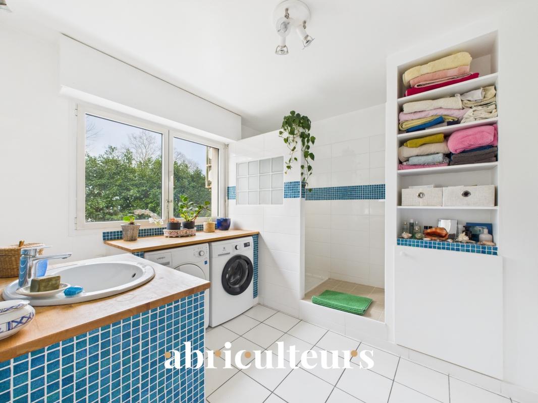 Bright laundry room with white tiles, blue mosaic accents, a washing machine, sink, and a window with potted plants on the sill.