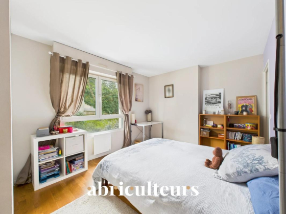 Bright bedroom with a large window and brown curtains, a neatly made bed with a teddy bear, white cubby storage, a small desk, and a wooden bookshelf filled with books and decor.