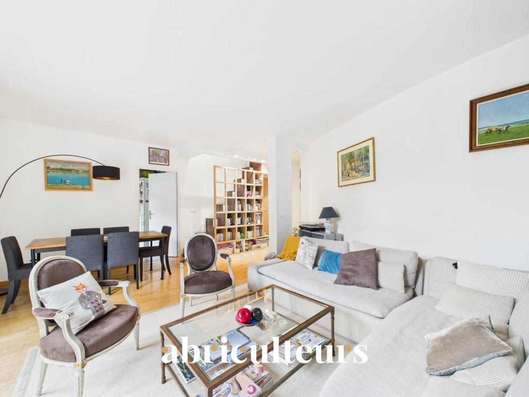Bright open-plan living and dining room with a beige sofa, matching armchairs, a glass coffee table, and a wooden dining table under natural light.