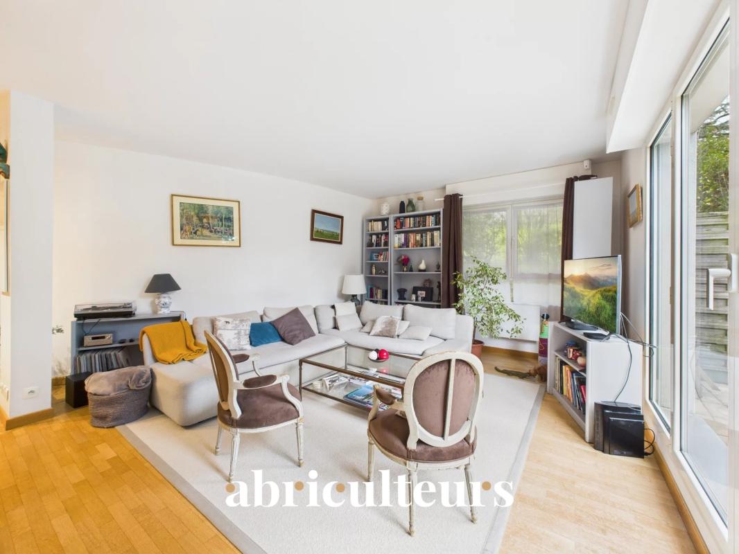 Bright living room with a pale sectional, two vintage chairs, a glass coffee table, and a large bookshelf by floor-to-ceiling windows.