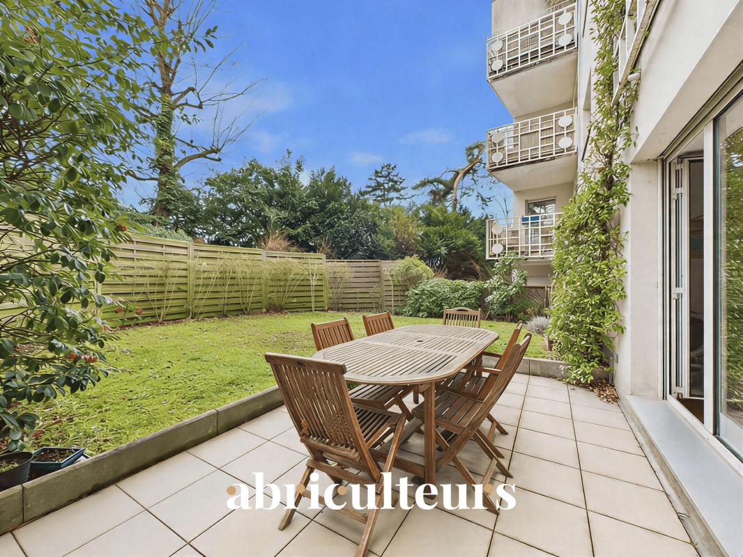 Outdoor patio with a wooden dining table and six chairs on a tiled terrace, overlooking a green lawn and residential building balconies in the background.