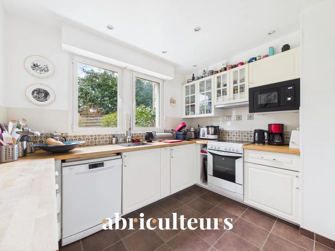 Bright white L-shaped kitchen with wooden countertops, dishwasher, oven, microwave, and glass-front cabinets under a large window over the sink; colorful jars line the upper cabinets and patterned backsplash shows at the counter edge.