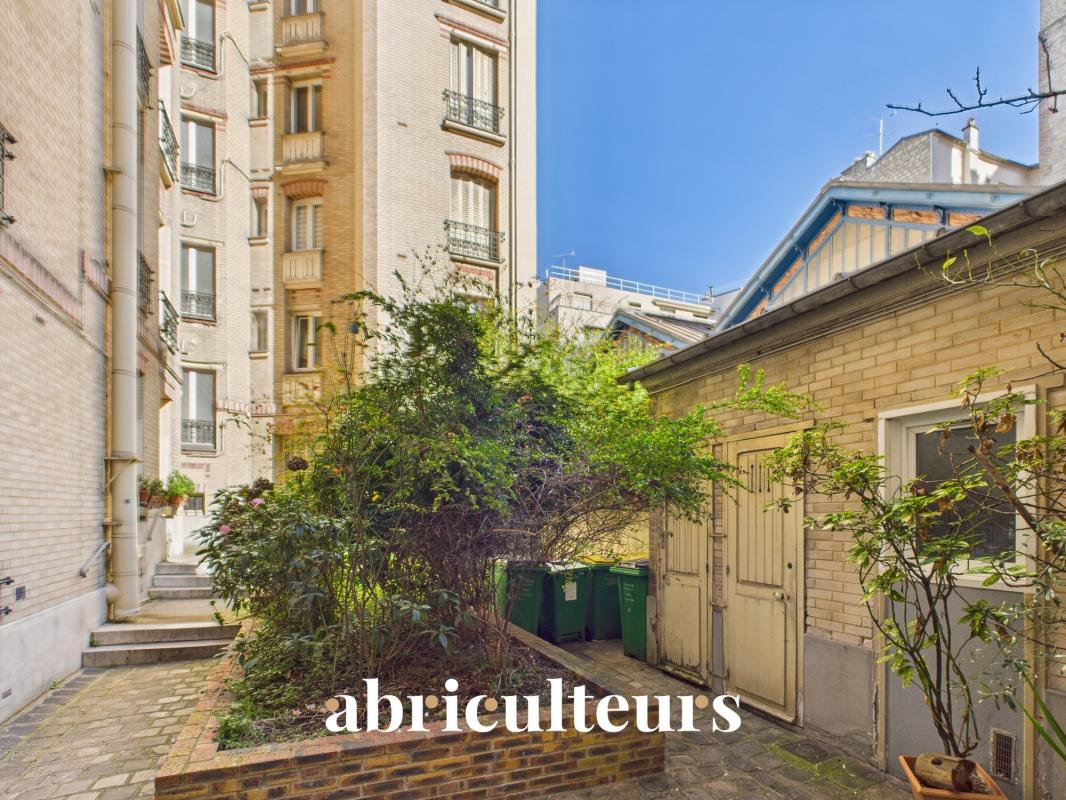 Urban courtyard between brick apartment buildings, with a large green shrub in a raised plant and several green waste bins along a brick path under a bright blue sky (text 'abriculteurs' overlaid).