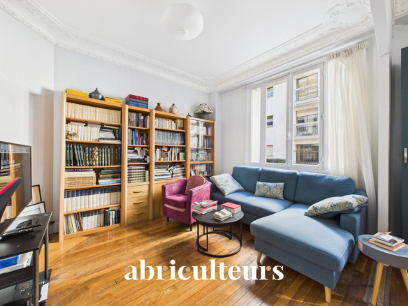 Bright living room with a blue L-shaped sofa, pink accent chair, and tall wooden bookcases filled with books, plus a round coffee table and large window letting in daylight; 'abriculteurs' logo appears at the bottom.
