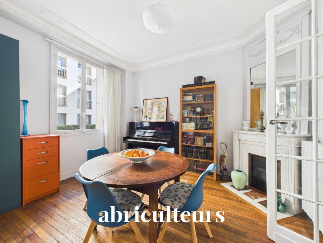 Bright dining room with a round wooden table, blue chairs, and a fruit bowl, beside a piano and bookcase.