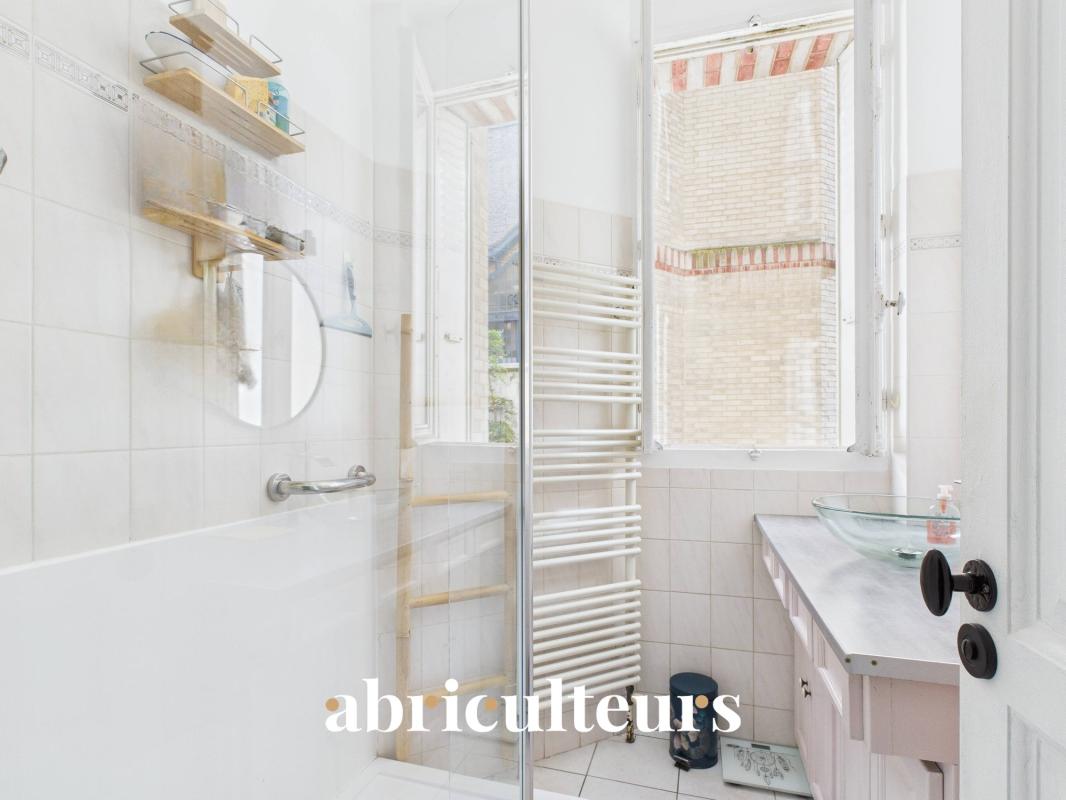 Bright bathroom with a glass shower enclosure, white tiled walls, and a wall-mounted towel warmer near a window.