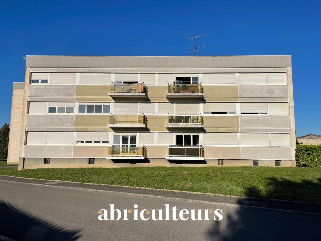 Four-story apartment building with beige-gray panels and small balconies against a blue sky; the word 'abriculteurs' appears along the street in the foreground.
