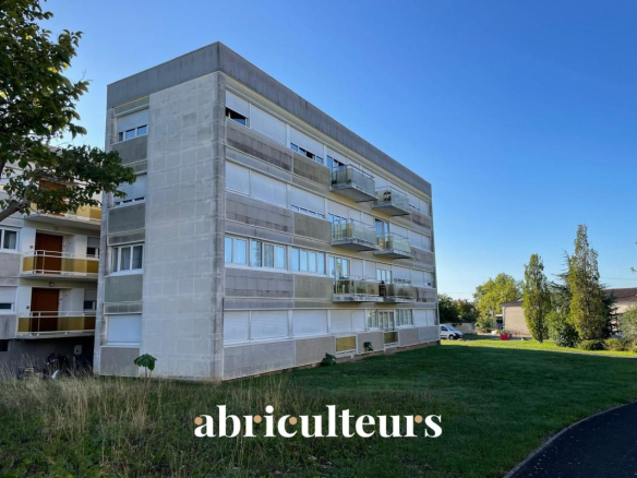 Four-story concrete apartment building with glass balconies under a clear blue sky, green lawn in front.