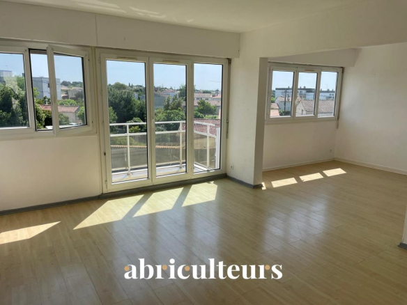 Bright, empty living room with large windows and a balcony door, light wood floors, and a city view outside.