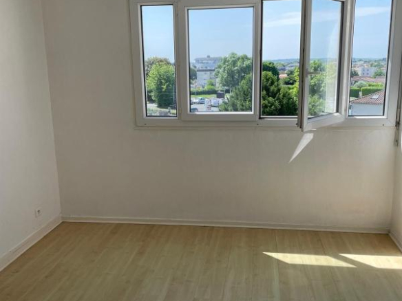 Bright empty room with white walls, light wood floor, and a large open window showing a tree-lined view outside.