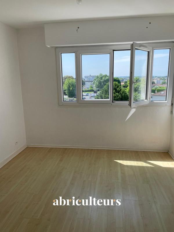 Bright empty room with white walls, light wood floor, and a large open window showing a tree-lined view outside.