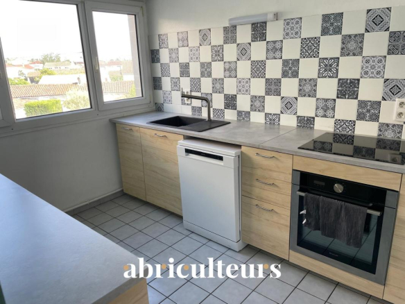 Bright L-shaped kitchen with light wood cabinets, tiled floor, and a blue‑and‑white patterned tile backsplash, a large window, stainless steel sink, dishwasher, and built‑in oven with an induction hob across from it.