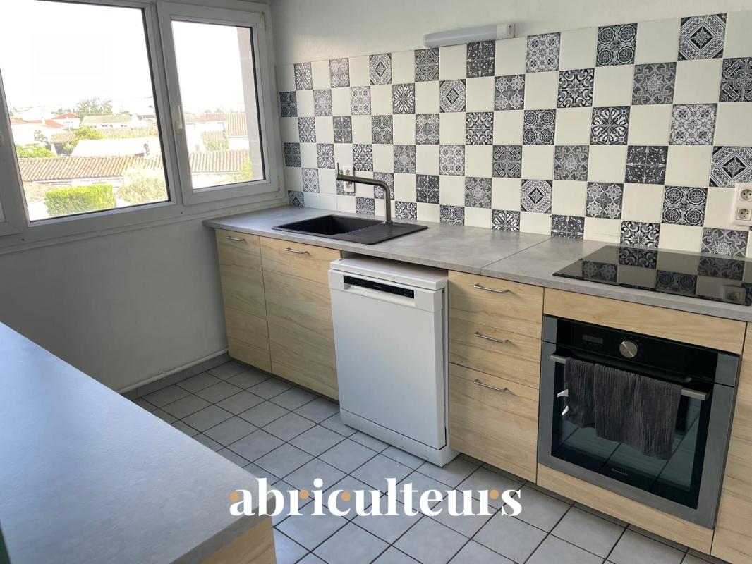 Bright L-shaped kitchen with light wood cabinets, tiled floor, and a blue‑and‑white patterned tile backsplash, a large window, stainless steel sink, dishwasher, and built‑in oven with an induction hob across from it.