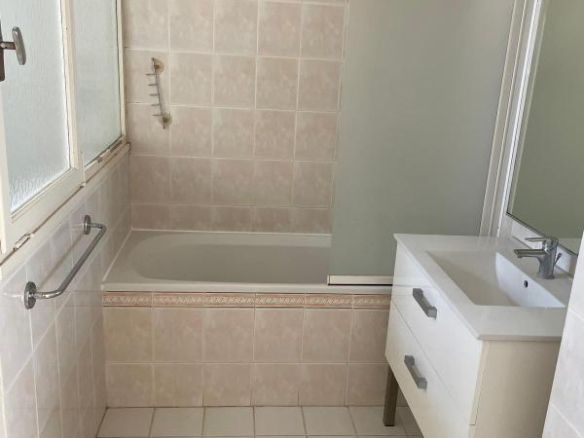 Bathroom with a built-in bathtub, frosted glass shower screen, white vanity, and beige tiled walls/flooring with a window on the left.