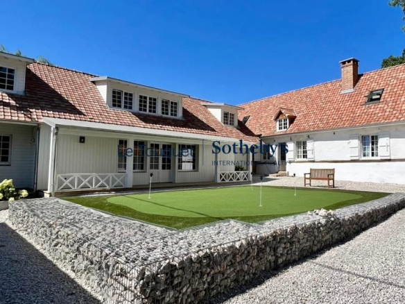 Exterior view of a white house with a red tile roof, dormer windows, and a gravel yard surrounding a small putting green with flags; Sotheby’s watermark across the image.