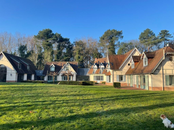 Row of cream-colored Tudor-style houses with brown roofs along a grassy lawn under a blue sky; a small dog in the bottom right corner.