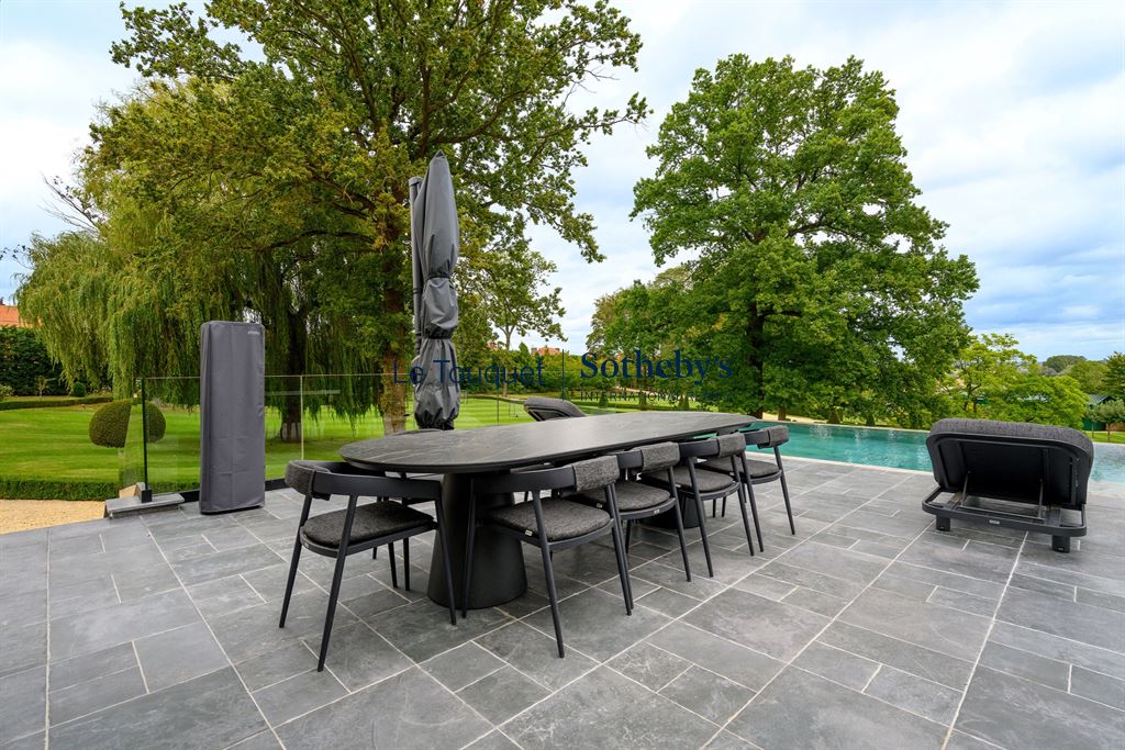 Terrasse d’un hôtel particulier à Mérignies Terrasse en pierre avec mobilier moderne, vue sur un parc verdoyant et une piscine extérieure.