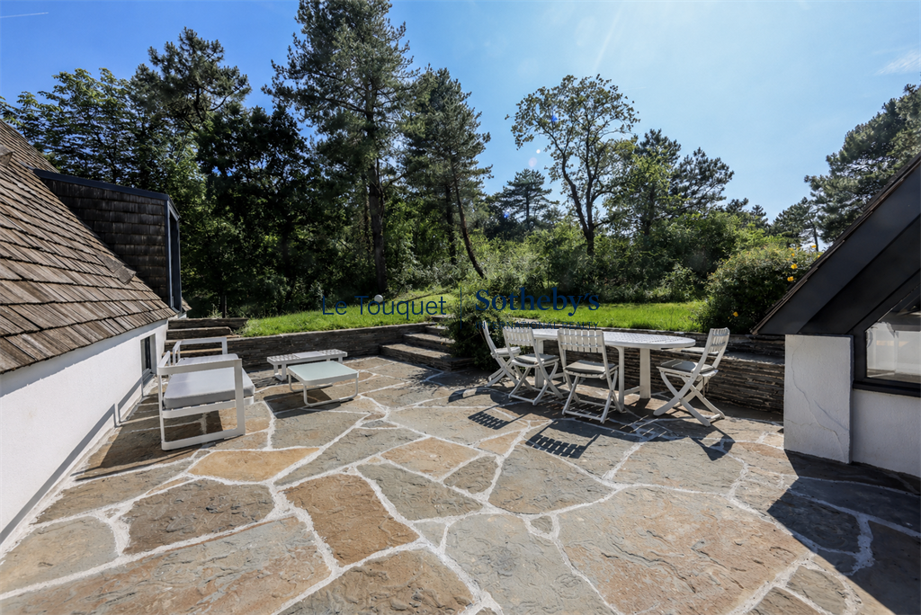 Terrasse de la villa d’architecte au Touquet Terrasse en pierre avec mobilier de jardin, entourée de pins maritimes et d'un paysage naturel au Touquet.